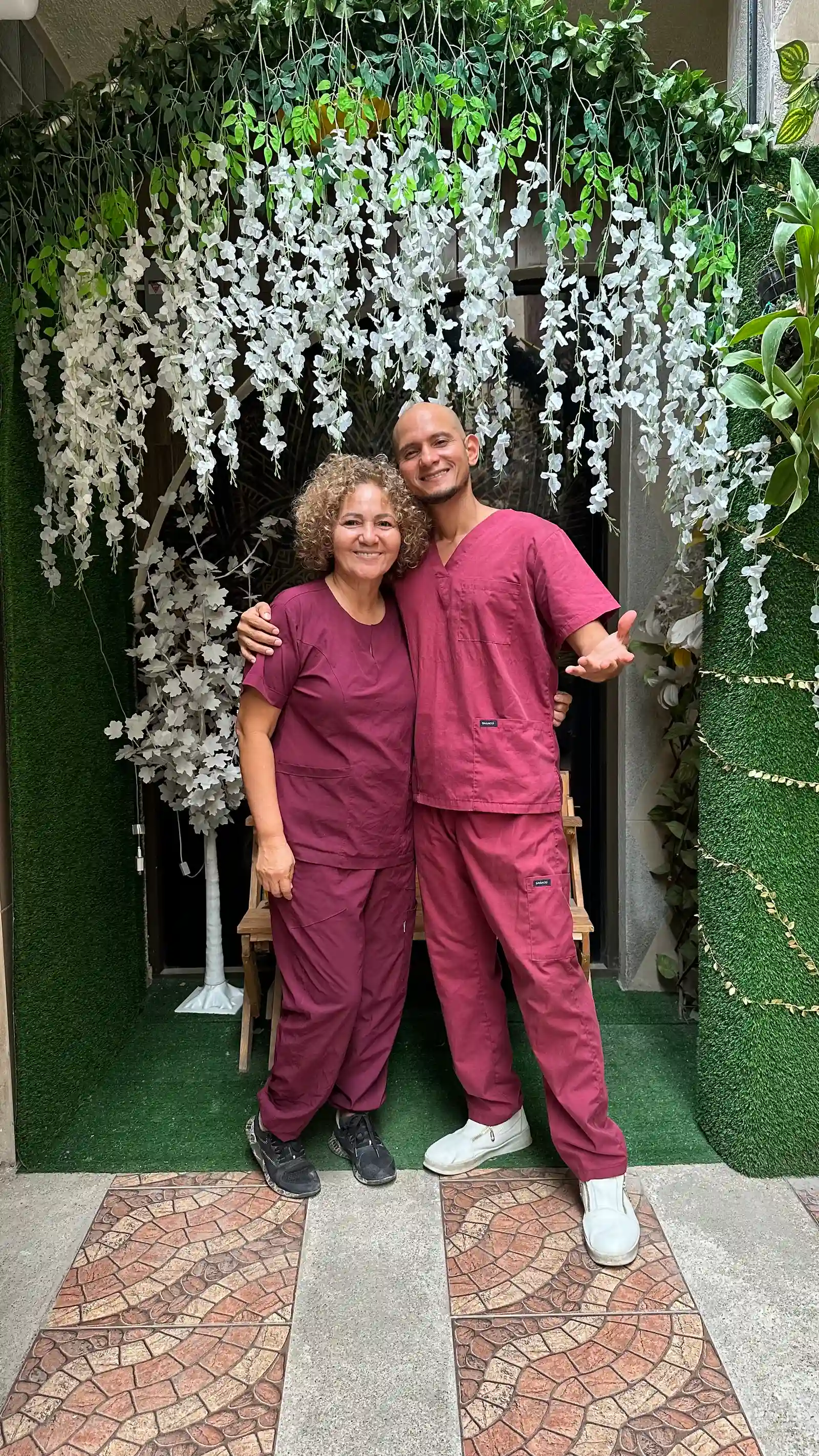Two SIRONA team members in burgundy uniforms pose beneath an arch of plants and white flowers.